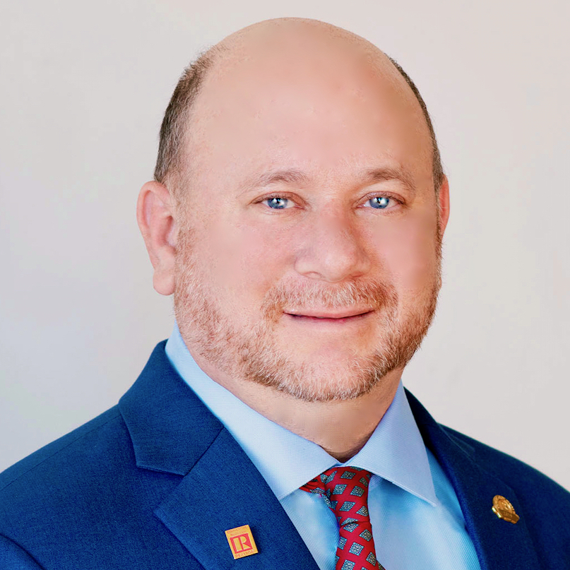 Russell wears a short beard and a patriotic blue suit with red tie. His REALTOR pin adorns his lapel.