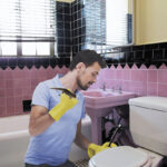 This image shows a young man in a blue short-sleeve polo shirt kneeling beside a closed toilet in a bathroom with pink and black tiles. He stares down at the toilet, casting a spell. He wears gloves on his hands. In his left hand, which rests next to the toilet lid, he holds a magic wand. Perched on his right hand, which is raised to shoulder height, there is a newt, also looking down at the toilet.