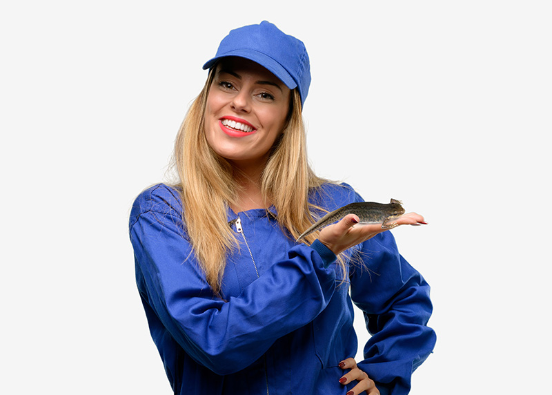 This image shows a blonde woman from the waist up. She wears blue plumber’s overalls, a blue cap, and is smiling. One hand is on her hip, the other is crossed in front of her shoulder, palm up. A newt is perched on her open palm.